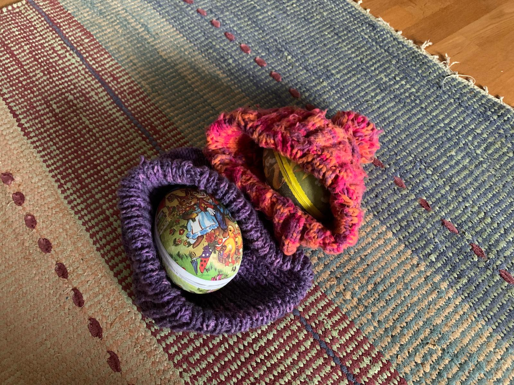 Två färggranna påskägg av papp instoppade i varsin färggran stickad mössa ligger på en trasmatta.